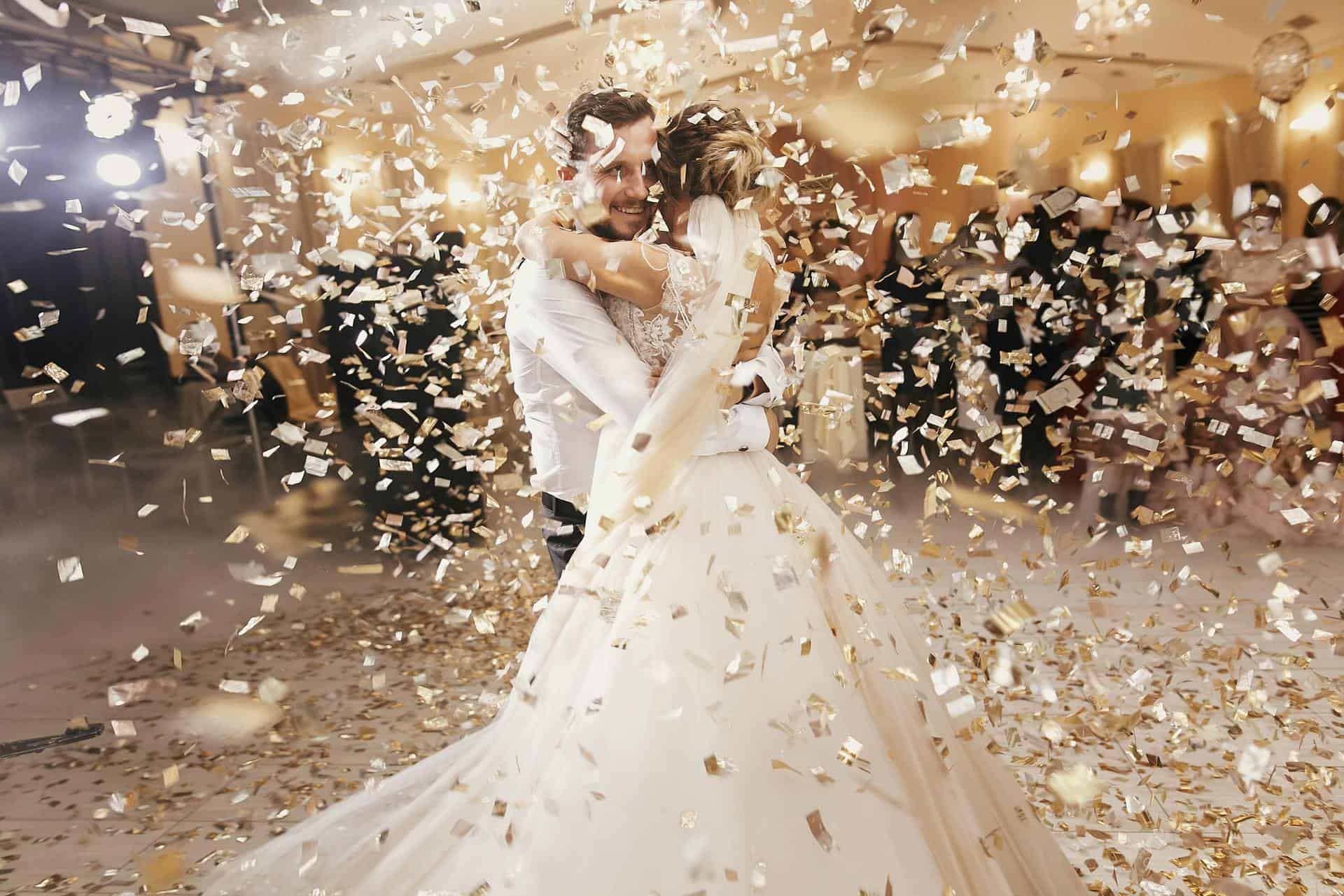 Gorgeous bride and stylish groom dancing under golden confetti at wedding reception